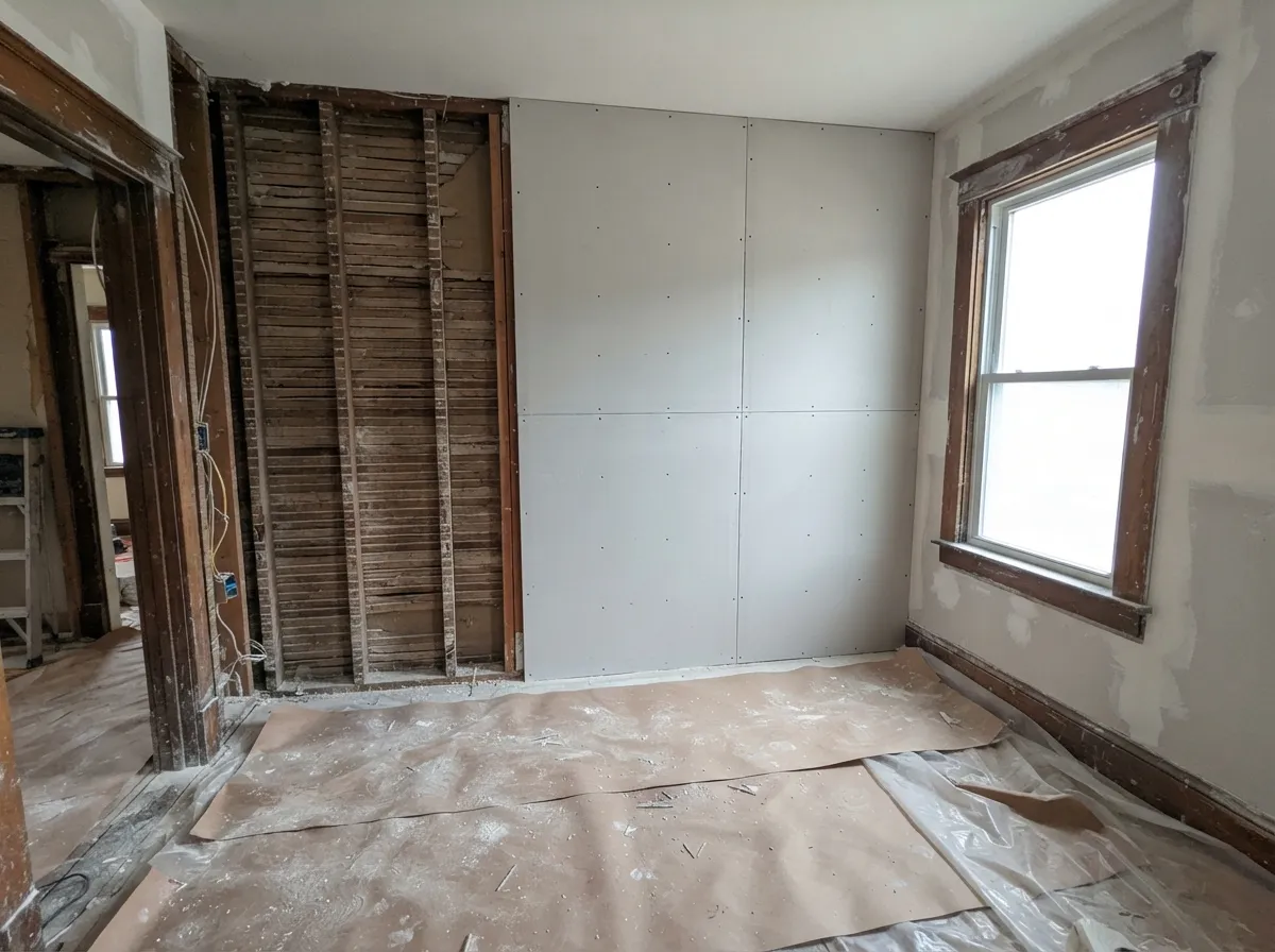 Plaster-to-drywall conversion in progress in an older house: new drywall screwed to one wall beside a section still open to the old lath, original wood window trim in place