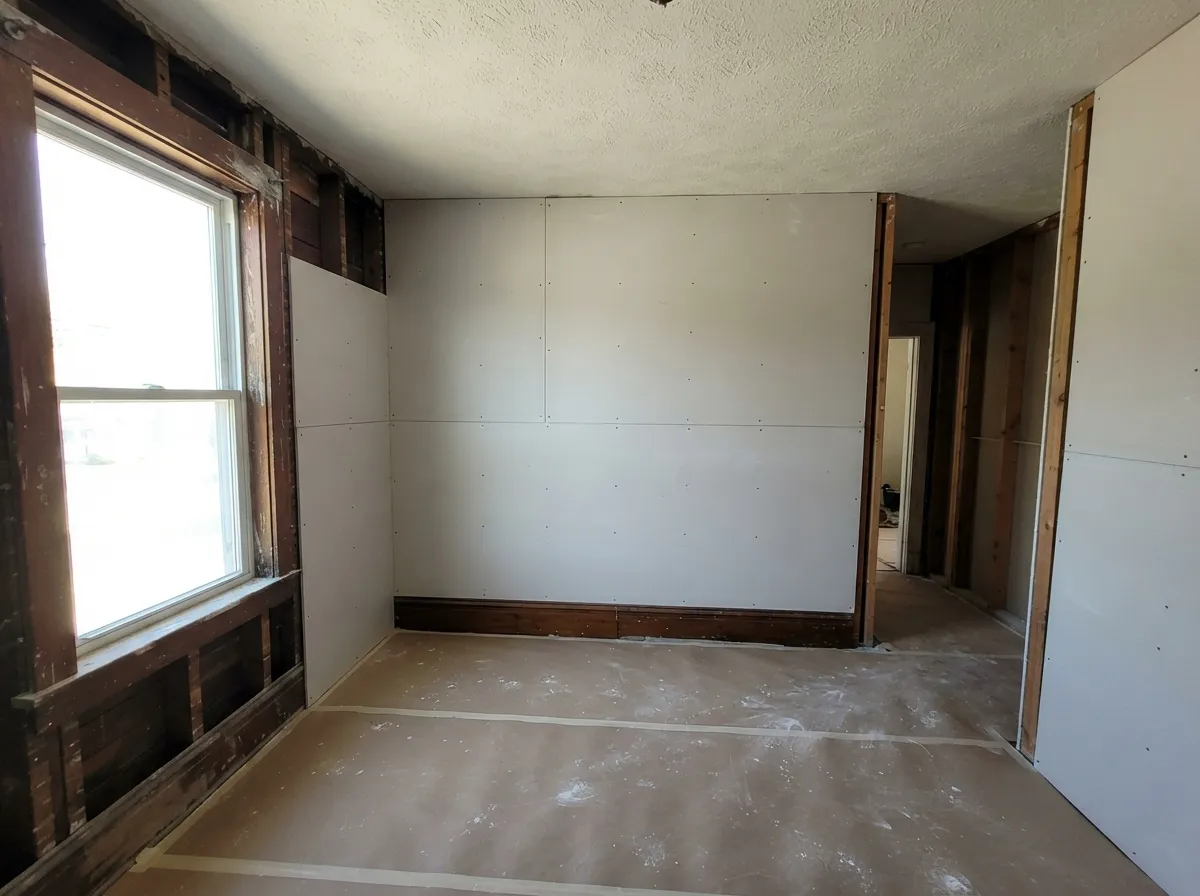 Older home mid-conversion: new drywall hung on the old studs where the plaster came out, original dark wood window trim and baseboard still in place