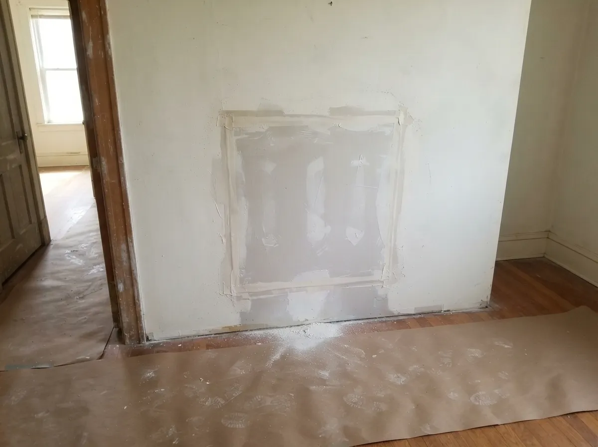 Square drywall patch in a wall beside a doorway, seams taped and first-coated, with paper protecting the wood floor